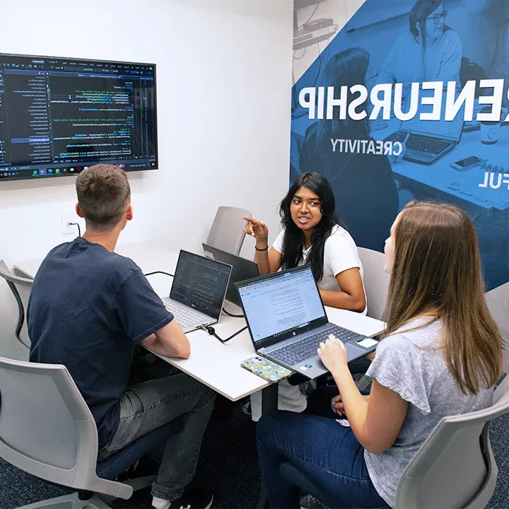 Three students gathered at table with laptops looking at computer code on a TV screen.